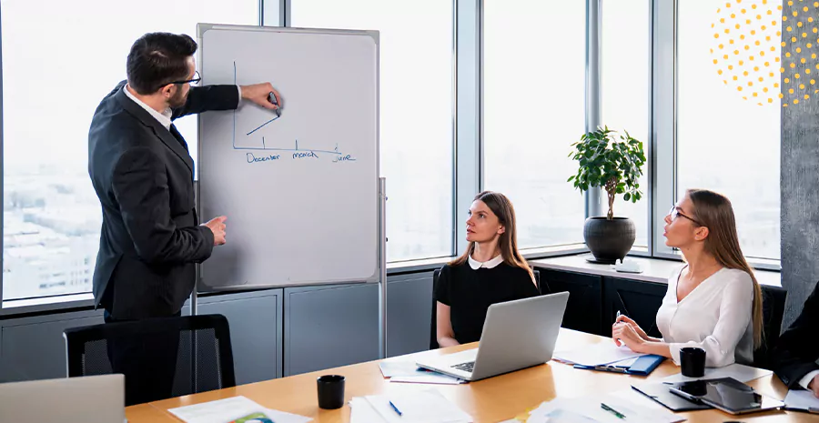 imagem de duas mulheres sentadas olhando para um homem em pé na frente delas apontando para um quadro branco