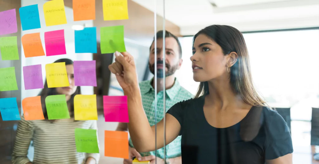 três pessoas, seno duas mulheres e um homem, olhando para um quadro com post-it