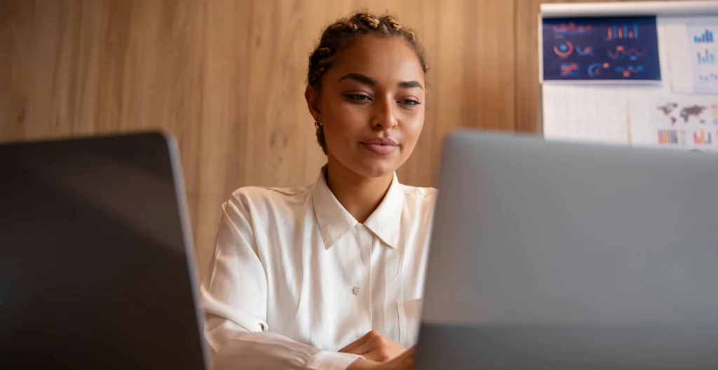 Mulher concentrada trabalhando em laptop, gráficos e estatísticas em painel ao fundo