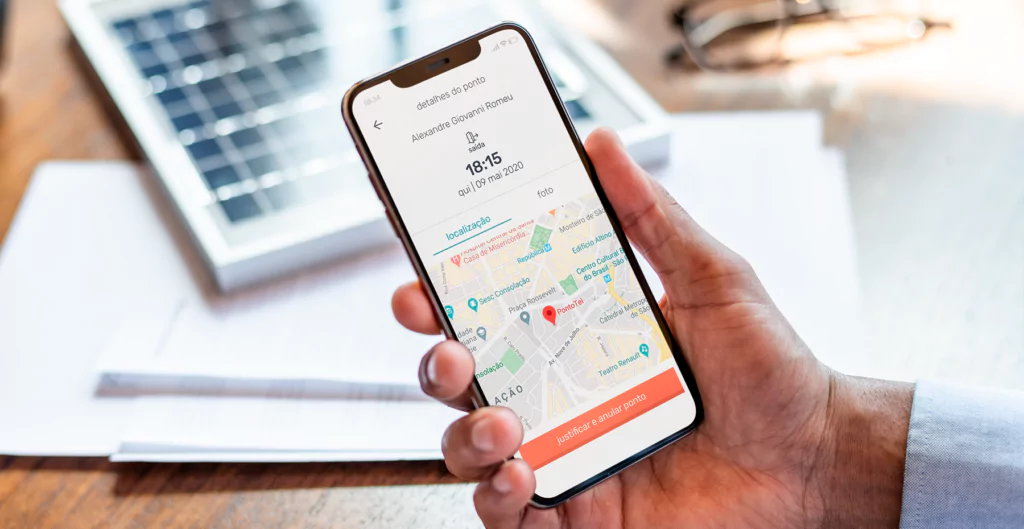 Mão segurando celular com app de controle de ponto e localização exibida.