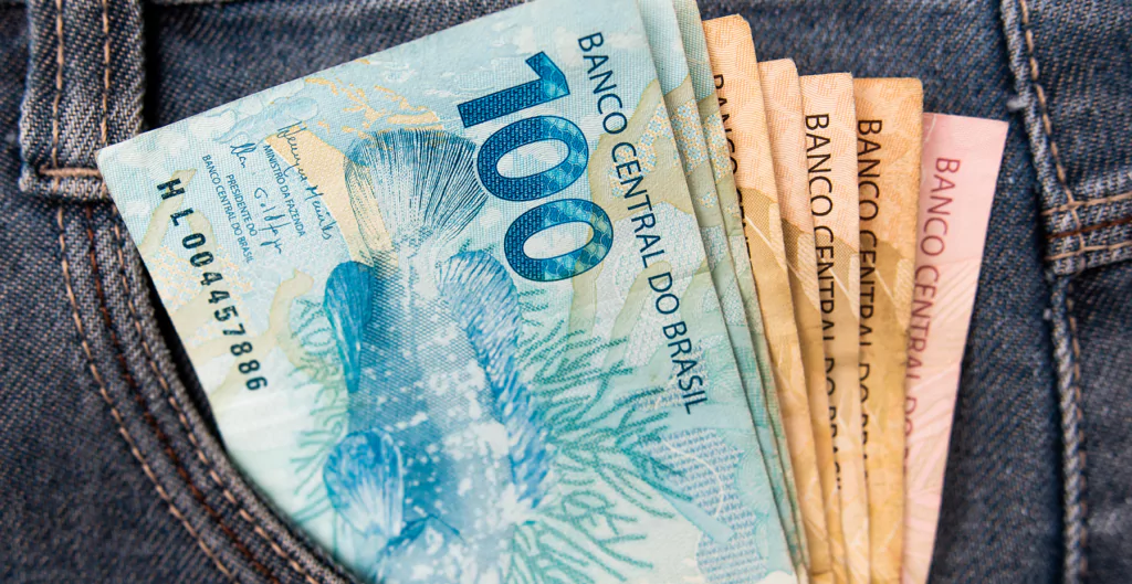 Notas de real no bolso de calça jeans, ilustrando recebimento de benefícios monetários.
