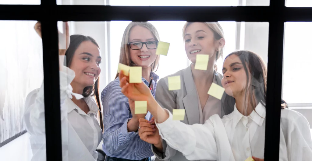 Quatro profissionais de mulher de negócios colaborando e planejando estratégias em uma reunião com post-its na parede, simbolizando criatividade e trabalho em equipe.