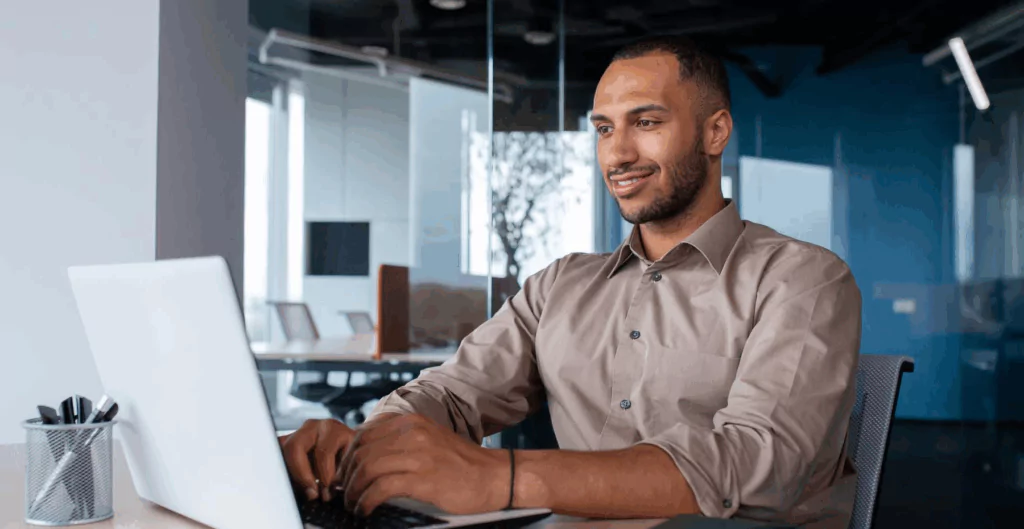 Homem sorridente usando computador portátil no escritório, ambiente corporativo moderno e bem iluminado, trabalhador confiável e atualizado.