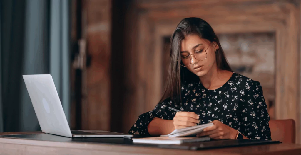 Mulher estudando com laptop e caderno em um ambiente aconchegante, indicando foco e dedicação ao estudo.