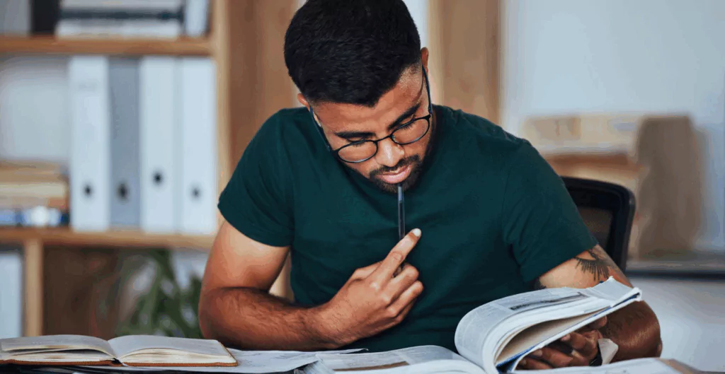 Homem estudando com livros e caderno, pensando e refletindo, em ambiente de escritório.