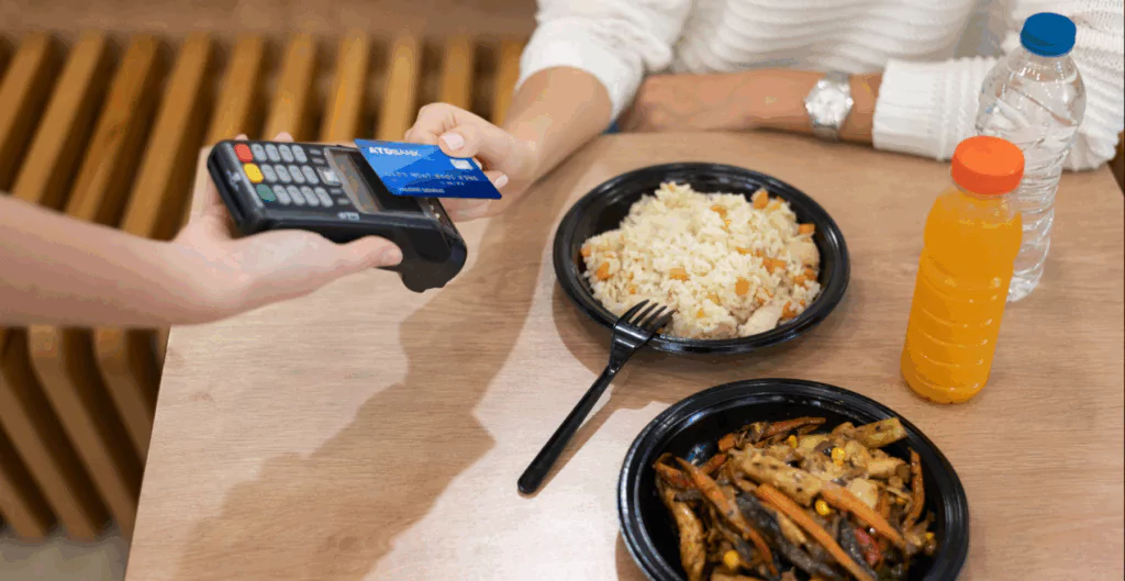 Pessoa pagando a conta em restaurante com cartão de crédito, mesas com pratos de comida, como arroz e legumes, e bebidas.