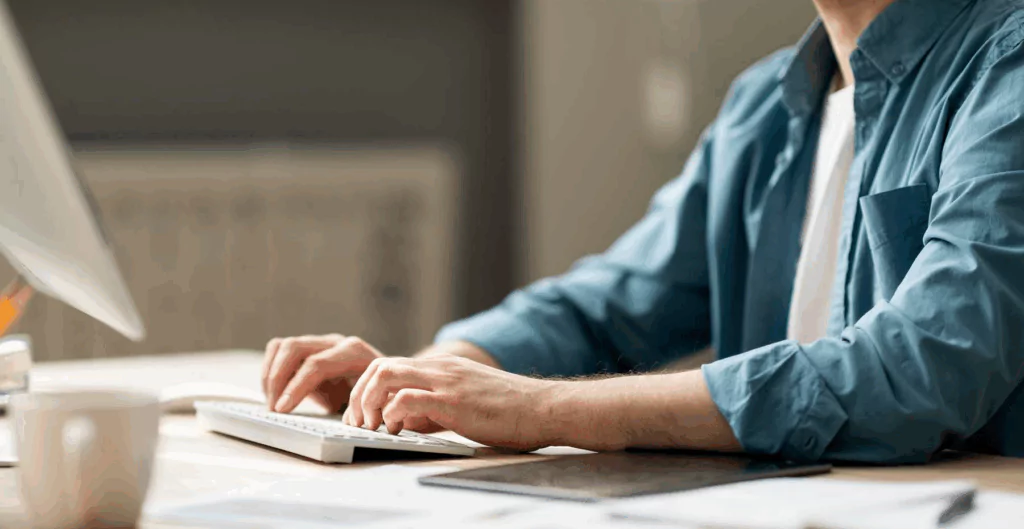 Homem trabalhando em computador na mesa de escritório, com foco nas mãos e sobre a mesa de trabalho, em um ambiente de escritório bem iluminado.