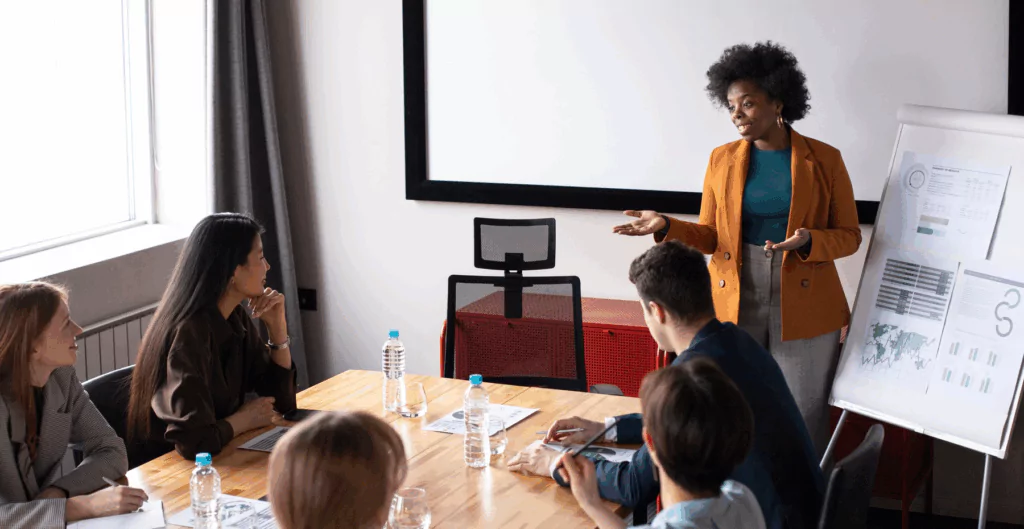 Professora ministrando aula de negócios para um grupo de estudantes em sala de aula moderna, com quadros de apresentação ao fundo e garrafas de água sobre a mesa.