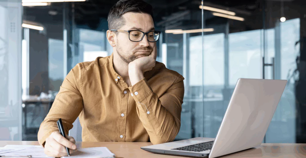 Homem pensativo usando laptop e caneta em escritório moderno, expressão de reflexão ou dúvida enquanto trabalha no computador.