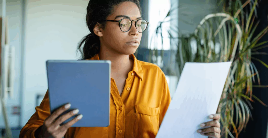 Jovem profissional analisando documentos e usando tablet em ambiente de escritório moderno, com foco em trabalho, leitura e organização.